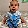 Carlos is registered to the contest to win money with this photo: baby, child, plush_toy, blue_toy, bedspread, pattern, indoor, cute, infant, smiling, sitting, hand, face, eyes, clothing, toy, soft_toy, portrait, person, young