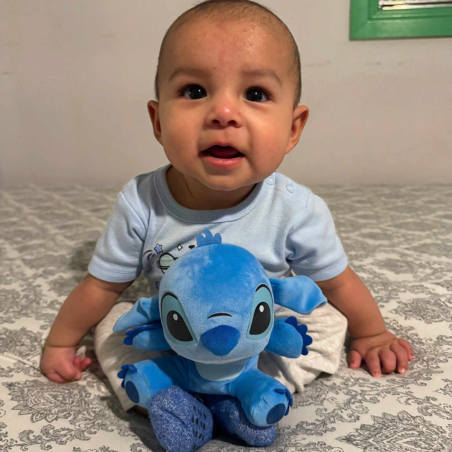 Carlos is registered to the contest to win money with this photo: baby, child, plush_toy, blue_toy, bedspread, pattern, indoor, cute, infant, smiling, sitting, hand, face, eyes, clothing, toy, soft_toy, portrait, person, young