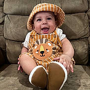 Carlos joined the competition — help win amazing prizes! baby, child, smiling, hat, overalls, lion_design, striped_clothing, sitting, couch, brown_couch, happy, cute, indoor, footwear, infant, person, portrait, clothing, headwear, baby_fashion