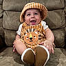 baby, child, smiling, hat, overalls, lion_design, striped_clothing, sitting, couch, brown_couch, happy, cute, indoor, footwear, infant, person, portrait, clothing, headwear, baby_fashion