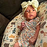animal_print, baby, bow_headband, child, comfort, cozy, curious_expression, cushion, cute, floral_dress, indoors, infant, lying_down, newborn, person, portrait, small_hands, soft_lighting, tender, woodland_animals