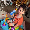 toddler, child, toy, activity_seat, bottle, orange_shirt, indoor, carpet, living_room, chair, table, curtains, furniture, playmat, expression, person, hair, floor, plastic_toy, casual_clothing
