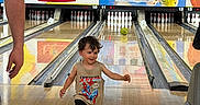 Elias is registered to the contest to win money with this photo: adult, american_flag, bowling_alley, bowling_ball, bowling_pins, child, colorful, flag, fun, hand, indoor, lane, marine_flag, recreation, running, smiling, spiderman, tank_top, toddler, wood_floor