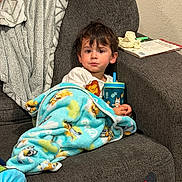 Elias is registered to the contest to win money with this photo: blanket, book, casual, child, comfort, couch, cozy, expression, footwear, furniture, home, indoor, person, relaxing, sippy_cup, sitting, socks, sofa, toddler, wall