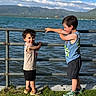 child, boy, outdoor, lake, water, fence, grass, mountain, sky, cloud, summer, casual_clothing, shorts, sneakers, smile, friendship, nature, daylight, playful, portrait