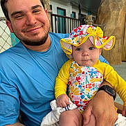 Leilani is registered to the contest to win money with this photo: baby, man, child, smiling, holding, colorful_clothing, sun_hat, towel, outdoor, casual, fence, watch, portrait, happy, baby_swimsuit, curious, seated, people, daylight, family