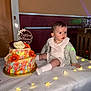 baby, child, cake, table, flowers, string_lights, indoors, celebration, baptism, white_clothing, cardigan, decorations, flower_cake, light, face, sitting, cute, party, event, infant