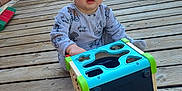 Nathanaël a rejoint le concours — aidez-le/la à gagner de superbes lots ! toddler, child, toy, shape_sorter, wooden_deck, brick_wall, outdoor, sunlight, greenery, hose, clothing, curious, sitting, playtime, baby, person, face, expression, casual, daytime