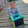 toddler, child, toy, shape_sorter, wooden_deck, brick_wall, outdoor, sunlight, greenery, hose, clothing, curious, sitting, playtime, baby, person, face, expression, casual, daytime