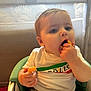 Mallory joined the competition — help win amazing prizes! baby, child, high_chair, snack, cheese_puffs, eating, white_shirt, blue_eyes, indoor, cute, chubby_cheeks, hand, face, mouth_open, green_chair, feeding, toddler, person, casual_clothing, happy