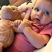 Mallory is registered to the contest to win money with this photo: baby, plush_toy, pink_clothing, pillow, soft_toy, indoor, child, cute, reclining, curious, wide_eyes, cuddly, comfort, person, infant, stuffed_animal, toddler, furniture, relaxed, closeup