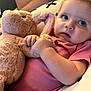 baby, plush_toy, pink_clothing, pillow, soft_toy, indoor, child, cute, reclining, curious, wide_eyes, cuddly, comfort, person, infant, stuffed_animal, toddler, furniture, relaxed, closeup