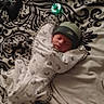 newborn, baby, sleeping, swaddle, hat, pacifier, bed, blanket, patterned_bedspread, controller, gaming, resting, cozy, infant, indoor, child, peaceful, soft_lighting, portrait, cute