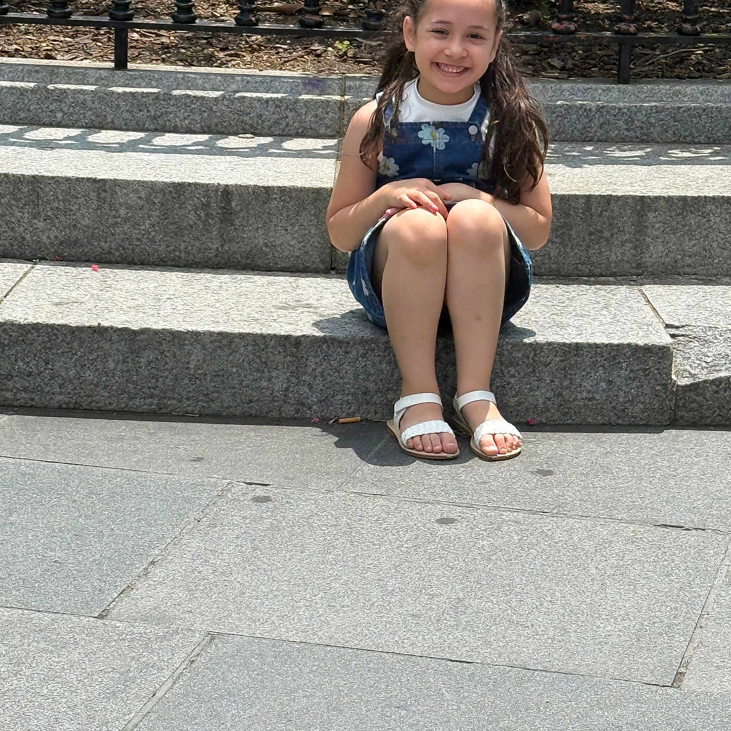 Sarah is registered to the contest to win money with this photo: child, girl, smile, sitting, steps, outdoor, sunlight, happy, braided_hair, dress, sandals, stone, pavement, fence, nature, daytime, casual, portrait, person, urban