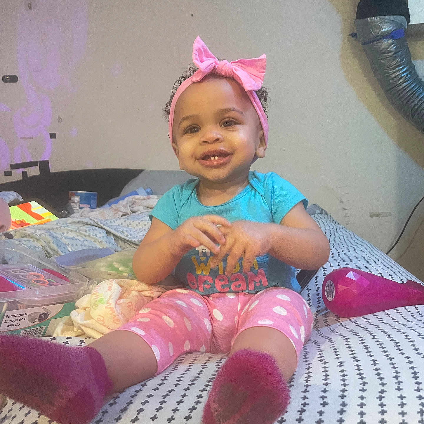 Raeven is registered to the contest to win money with this photo: baby, bed, blanket, child, clothing, furniture, happy, headband, indoor, pattern, person, pink_bow, polka_dot_pants, room, smiling, socks, storage_box, toddler, toys, ventilation_duct