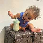 Raeven joined the competition — help win amazing prizes! toddler, child, baby_bottle, curly_hair, blue_clothing, woven_box, indoor, foot, hand, milk, curious, expression, furniture, storage_box, vent, wall, sitting, playful, person, closeup