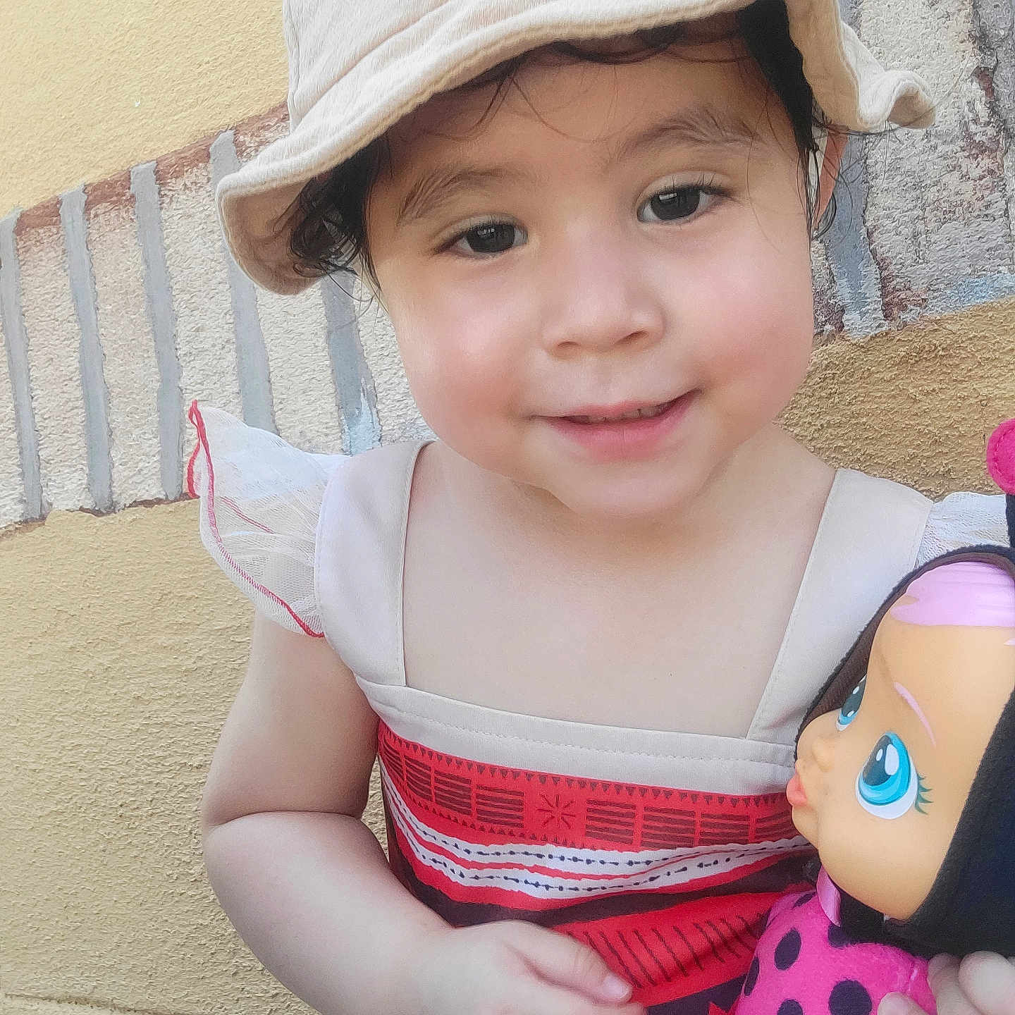 Meilyn is registered to the contest to win money with this photo: child, toddler, smiling, portrait, outdoor, sun_hat, dress, doll, toy, cuddling, face, eyes, hair, happy, wall, textured_background, close_up, standing, playtime, photography