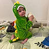 baby, bed, blanket, child, cozy, curious, cute, dinosaur_onesie, foot, giraffe_print, green_costume, indoor, jungle_theme, playful, plush_toy, sitting, smiling, soft_toys, toddler, wall
