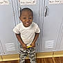 background, boy, camouflage_pants, cartoon_stickers, casual, child, clothing, expression, flooring, funny_face, holding_object, indoor, locker, person, playful, school, standing, white_sneakers, white_tshirt, wooden_floor