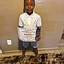 achievement, boy, cap, carpet, celebration, certificate, child, graduation, holding, indoor, kindergarten, person, portrait, proud, shirt, shoes, shorts, smile, standing, wall