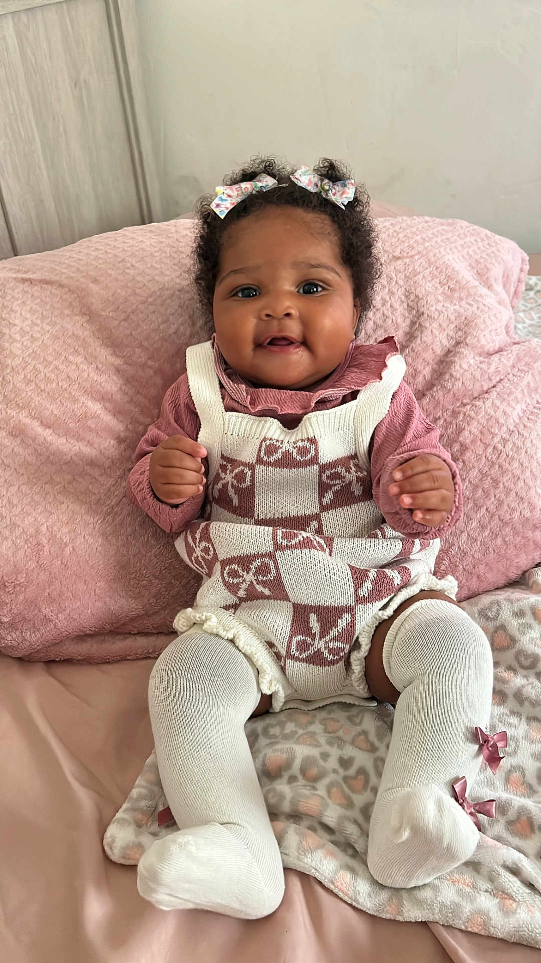 Genesis is registered to the contest to win money with this photo: baby, child, smiling, curly_hair, bows, knitwear, white_socks, blanket, pink, cute, indoor, portrait, happy, infant, soft_texture, cozy, sitting, clothing, face, hands