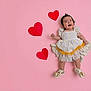 baby, child, smiling, dress, white_dress, yellow, hearts, pink_background, lying_down, happy, cute, footwear, shoes, celebration, adorable, infant, portrait, studio_photo, colorful, playful