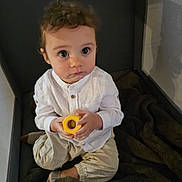 Lowen a rejoint le concours — aidez-le/la à gagner de superbes lots ! toddler, child, toy, playpen, blanket, white_shirt, beige_pants, curious, indoor, sitting, holding, wooden_toy, cross-legged, young_child, face, person, cute, brown_hair, large_eyes, soft_lighting