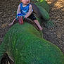 toddler, child, statue, crocodile, outdoor, daylight, green, playground, sippy_cup, blue_clothing, shorts, barefoot, woodchips, tree, sunlight, curious, toy, nature, park, summer