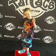Zayden is registered to the contest to win money with this photo: child, smiling, peace_sign, red_carpet, colorful_clothing, backdrop, logo, hashtag, event, footwear, standing, pose, fun, indoor, person, happy, shorts, tshirt, blue_shoes, carpet