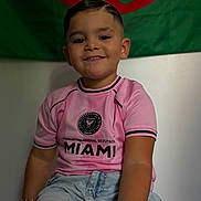 Waylon a rejoint le concours — aidez-le/la à gagner de superbes lots ! child, smiling, portrait, indoor, tshirt, pink_clothing, denim_shorts, short_hair, flag, green_background, red_pattern, happy, casual, sitting, person, cute, fashion, young_boy, sportswear, bracelet