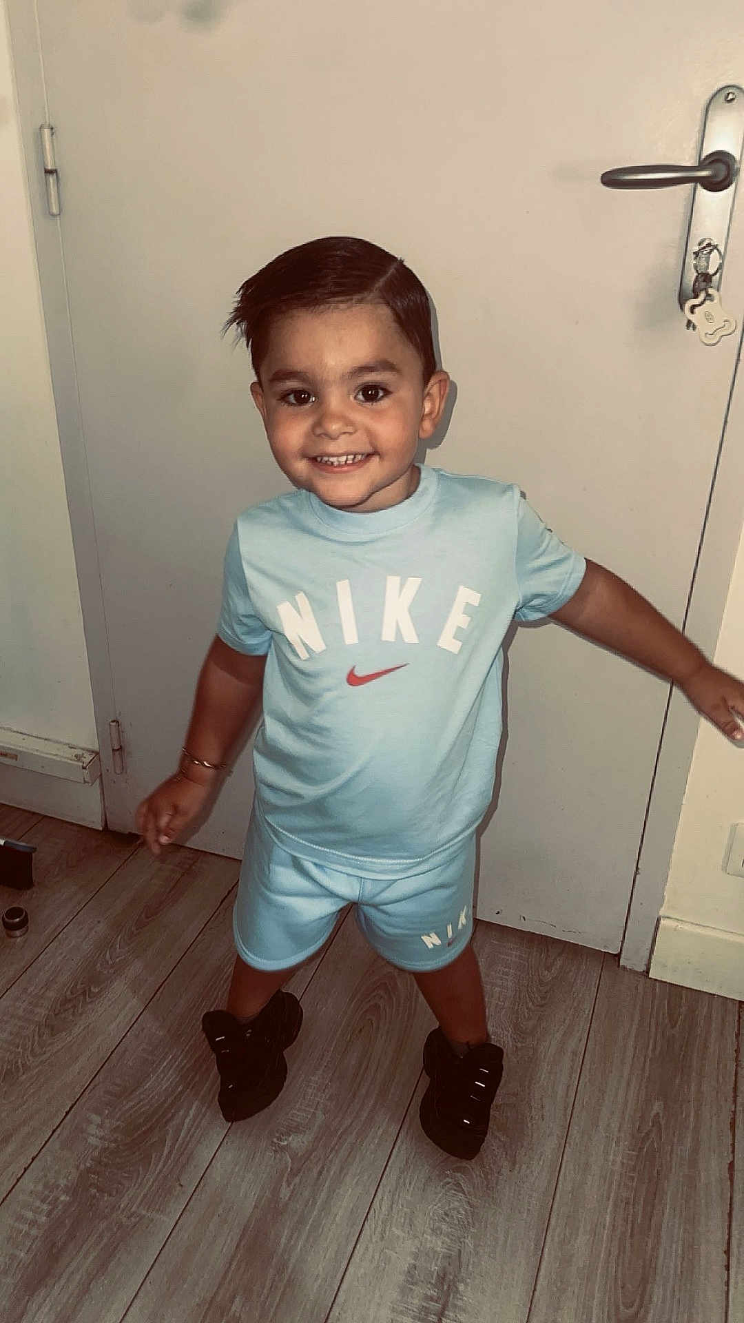 Waylon participe au concours pour gagner de l'argent avec cette photo : child, toddler, smiling, standing, indoors, door, keychain, floor, wooden_floor, clothing, nike, shorts, tshirt, black_shoes, hair, happy, person, pose, casual, home