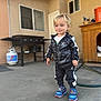 toddler, child, boy, smiling, standing, tracksuit, sneakers, outdoor, patio, concrete, propane_tank, grill, doghouse, window, hose, building, casual, daylight, cute, happy