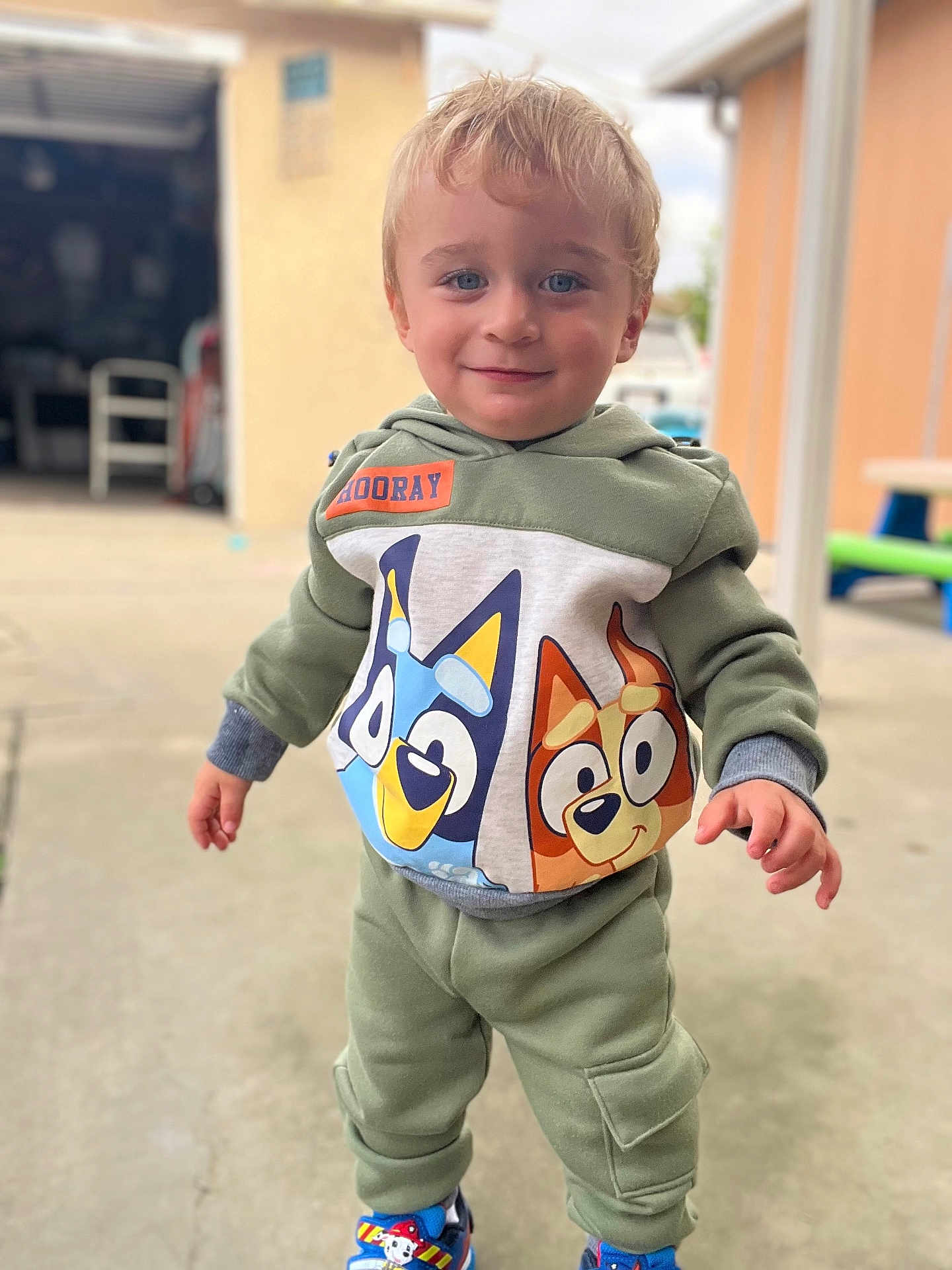 Atom is registered to the contest to win money with this photo: child, toddler, hoodie, cartoon, outdoor, smile, face, pants, shoes, concrete, playful, standing, person, blue_eyes, blond_hair, casual_clothing, daylight, background_blur, toy, happy