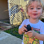 Atom is registered to the contest to win money with this photo: blonde, child, concrete, curly_hair, face, fence, garden_hose, grass, happy, holding, lattice, outdoor, person, playful, rock, smiling, sunlight, teenage_mutant_ninja_turtles, toddler, tshirt