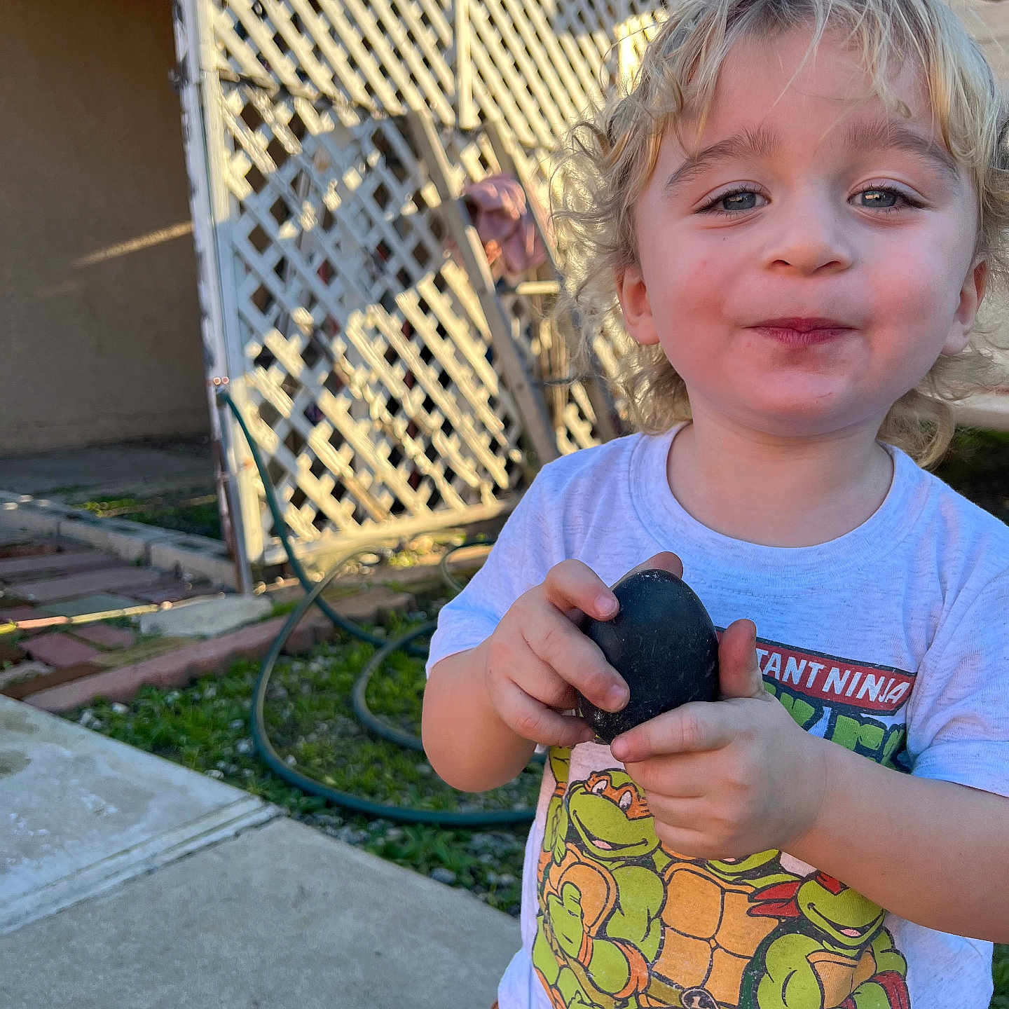 Atom is registered to the contest to win money with this photo: blonde, child, concrete, curly_hair, face, fence, garden_hose, grass, happy, holding, lattice, outdoor, person, playful, rock, smiling, sunlight, teenage_mutant_ninja_turtles, toddler, tshirt