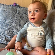 Atom is registered to the contest to win money with this photo: blue_eyes, candid, casual, child, couch, curious, curly_hair, diaper, food, indoor, light, long_sleeve_shirt, messy_face, patterned_cushion, pointing, portrait, sitting, snack_container, toddler, young_child