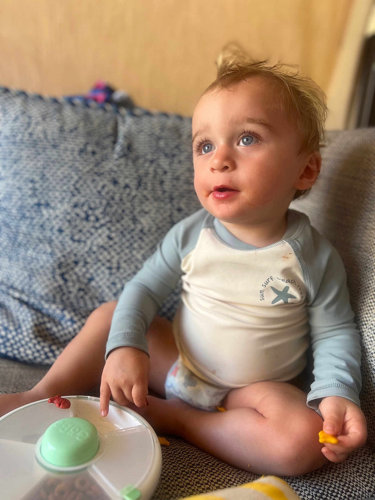 Atom is registered to the contest to win money with this photo: blue_eyes, candid, casual, child, couch, curious, curly_hair, diaper, food, indoor, light, long_sleeve_shirt, messy_face, patterned_cushion, pointing, portrait, sitting, snack_container, toddler, young_child