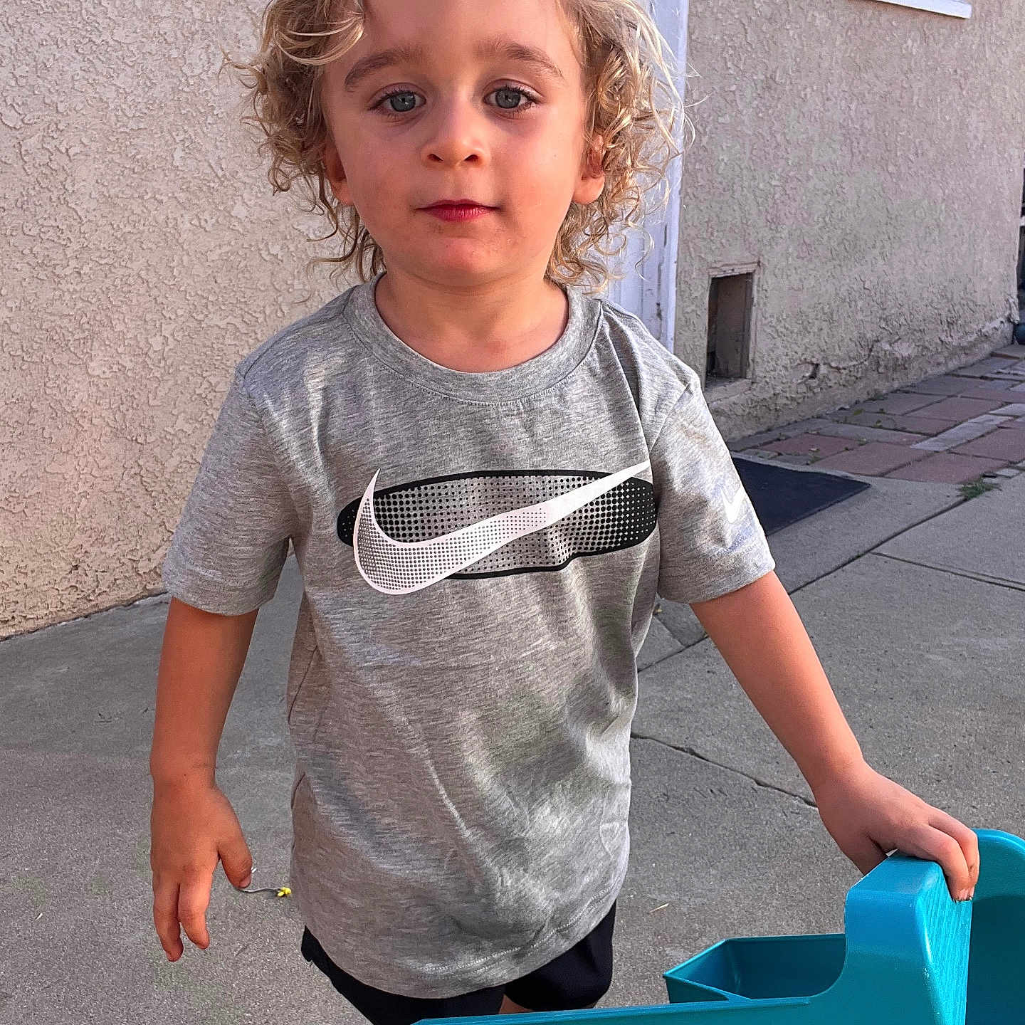 Atom joined the competition — help win amazing prizes! child, curly_hair, blonde, grey_tshirt, nike_logo, toy_truck, outdoor, concrete, sidewalk, wall, door, young_child, casual_clothing, portrait, playtime, sunlight, expression, person, blue_toy, summer