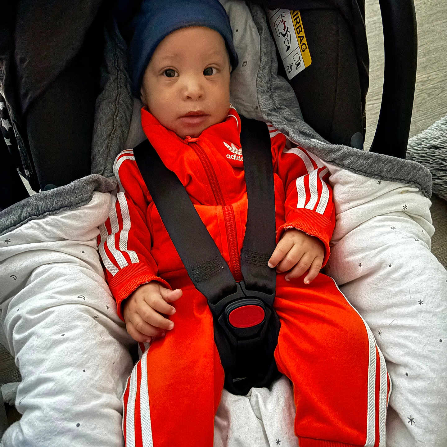 Laël a rejoint le concours — aidez-le/la à gagner de superbes lots ! baby, baby_clothes, blanket, blue_hat, car_seat, child, clothing, cushion, cute, face, footwear, indoor, infant, orange_tracksuit, person, safety, seated, small_hands, socks, straps
