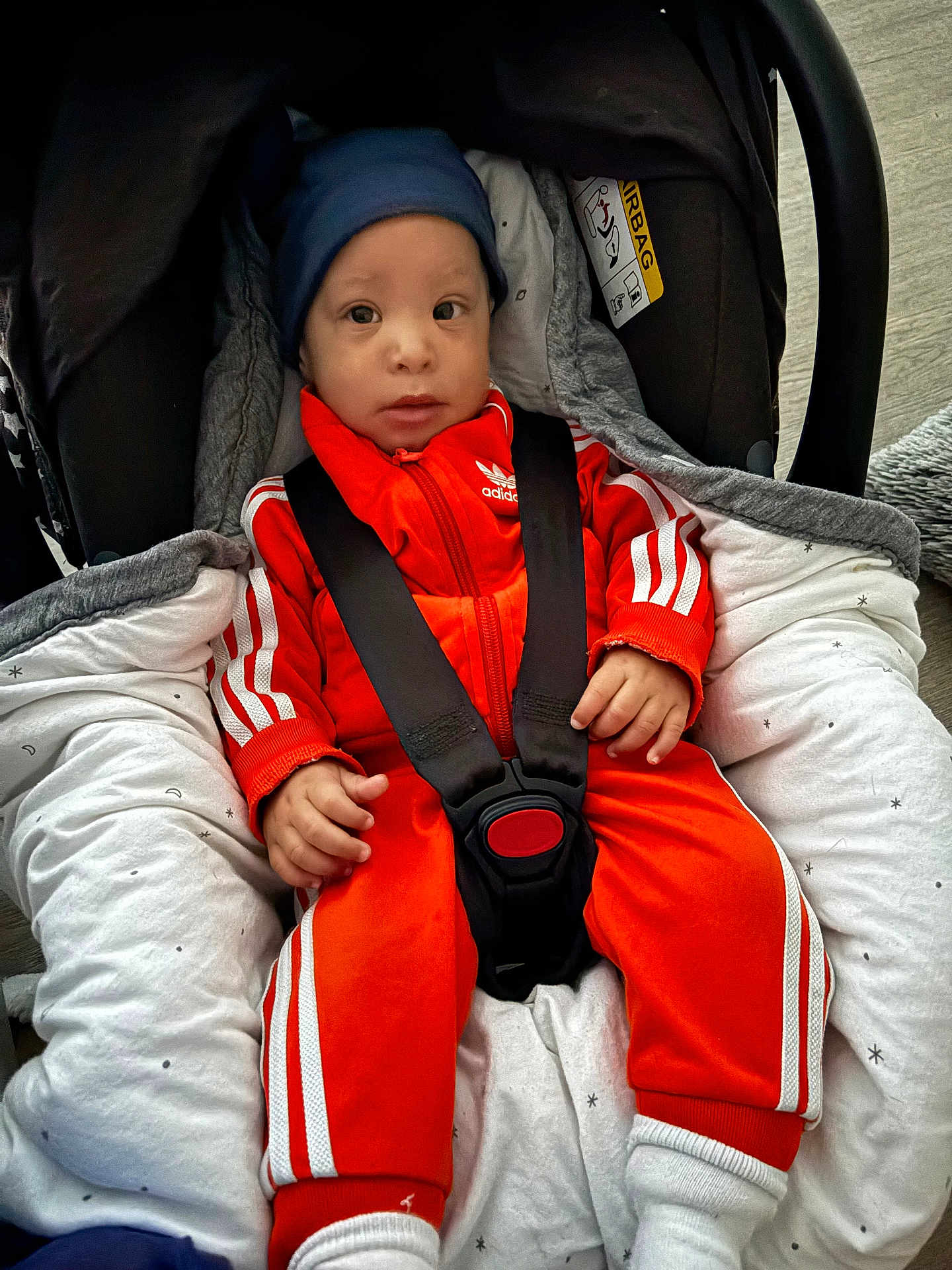 Laël a rejoint le concours — aidez-le/la à gagner de superbes lots ! baby, infant, car_seat, orange_tracksuit, blue_hat, straps, cushion, blanket, clothing, person, indoor, footwear, socks, baby_clothes, face, small_hands, seated, cute, child, safety