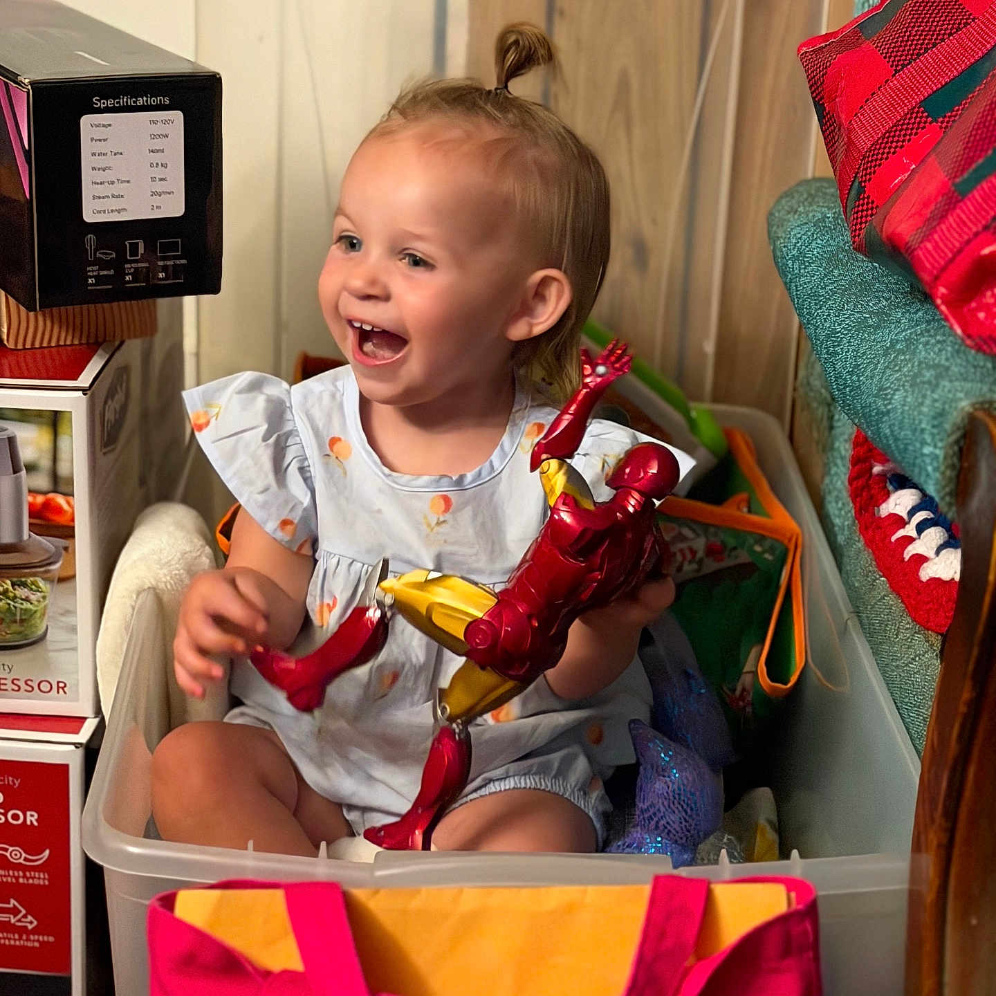 Remington is registered to the contest to win money with this photo: toddler, child, smiling, toy, action_figure, iron_man, plastic_bin, indoor, playful, happy, person, clothing, bag, home, furniture, floor, hair, cute, sitting, childhood