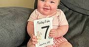 Aurelia joined the competition — help win amazing prizes! baby, infant, smiling, sitting, couch, milestone_card, pink_shirt, checkered_shorts, barefoot, cute, child, indoors, home, portrait, face, hands, feet, happy, young, person
