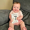 baby, infant, smiling, sitting, couch, milestone_card, pink_shirt, checkered_shorts, barefoot, cute, child, indoors, home, portrait, face, hands, feet, happy, young, person
