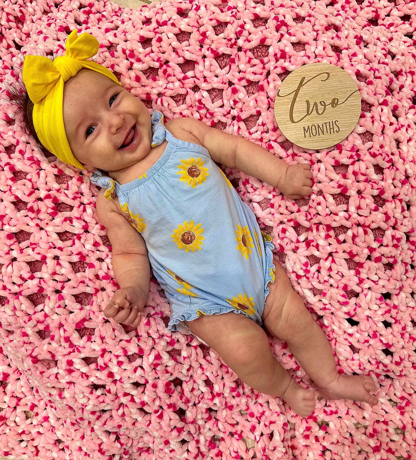 Zoe joined the competition — help win amazing prizes! baby, infant, smiling, yellow_headband, blue_onesie, sunflower_pattern, pink_blanket, wooden_sign, two_months, lying_down, happy, cute, child, portrait, indoors, soft_texture, headwear, clothing, skin, baby_girl