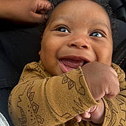 Carter is registered to the contest to win money with this photo: baby, smiling, infant, face, hand, brown_skin, curly_hair, clothing, dinosaur_pattern, closeup, joy, cute, happy, portrait, child, fist, seated, fabric, texture, expression
