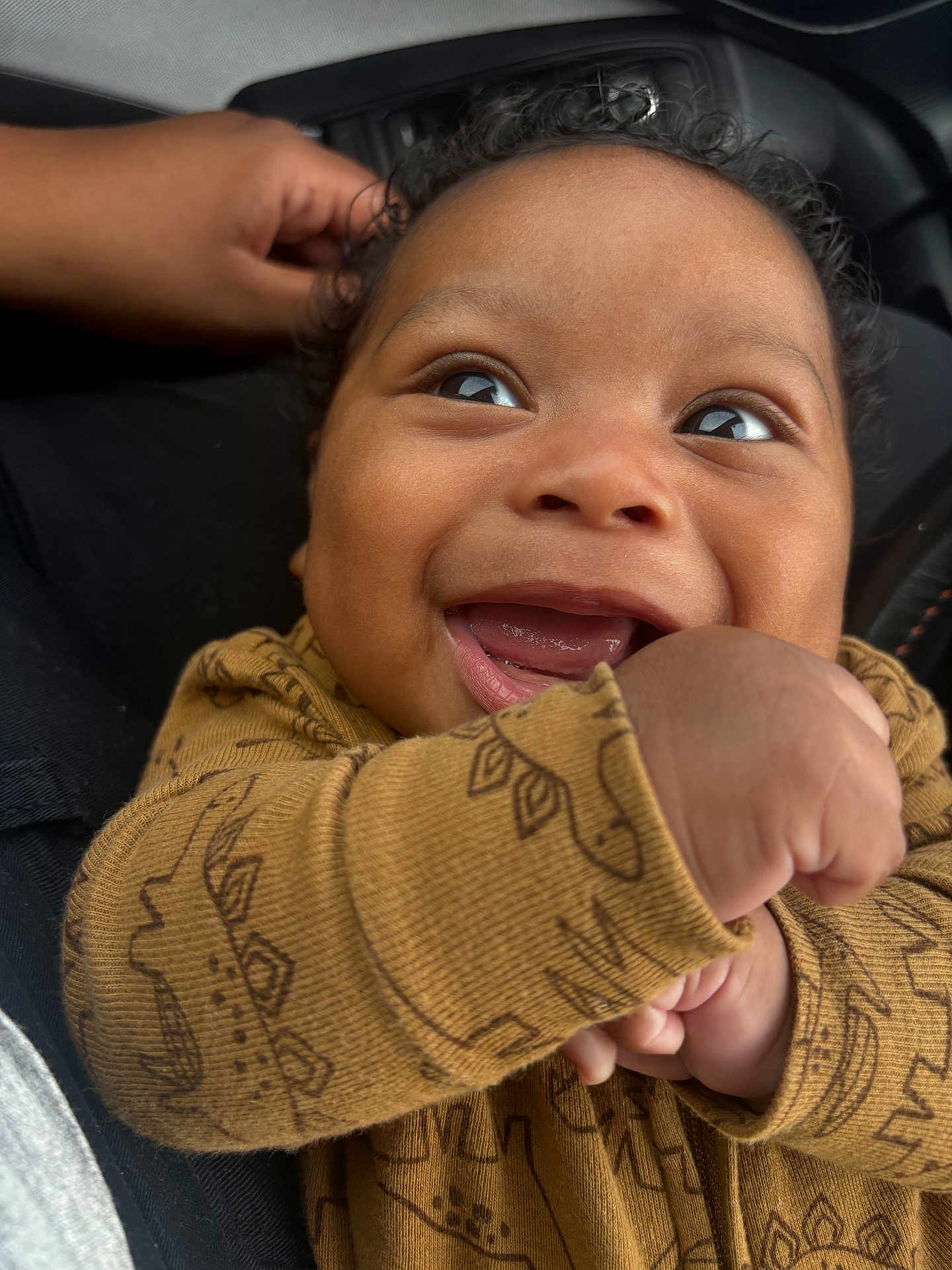 Carter is registered to the contest to win money with this photo: baby, smiling, infant, face, hand, brown_skin, curly_hair, clothing, dinosaur_pattern, closeup, joy, cute, happy, portrait, child, fist, seated, fabric, texture, expression