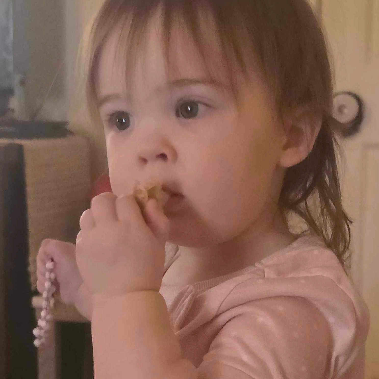 Charlotte is registered to the contest to win money with this photo: beads, bracelet, casual, child, closeup, curly_hair, cute, expression, face, hand, home, indoors, nibble, person, pink_clothing, portrait, snack, soft_lighting, toddler, young_child
