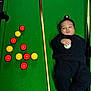 8_ball, baby, balanced_object, billiard_balls, black_clothing, casual, child, colorful_balls, cue_ball, curious_expression, funny_pose, game_room, green_felt, indoor, lying_down, playing, pool_cues, pool_table, recreation, sports