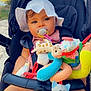 baby, baby_clothing, blue_eyes, cat_toy, child, cute, daylight, giraffe_toy, hat, infant, outdoor, pacifier, person, plush_toy, relaxing, seatbelt, smiling, soft_toys, stroller, sun_hat