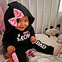 baby, black_clothing, child, cozy, crib, cute, hoodie, indoor, infant, person, pink_accents, pink_bow, playful, portrait, sitting, smiling, soft_toys, stuffed_animals, toddler, white_text
